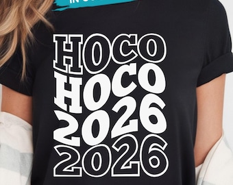 Op de afbeelding: Een zwart t-shirt met korte mouwen en witte grafische tekst die HOCO HOCO 2026 2026 leest in een gestapeld, vet en golvend lettertype. Het ontwerp is gecentreerd op de borst en een kleine banner bovenaan toont de tekst IN COLORS met twee cirkels.