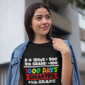K&ouml;nnte beinhalten: Ein schwarzes T-Shirt mit dem Text "K-4: 180x5 = 900, 5th GRADE: +100, 1000 DAYS SMARTER, #5th GRADE" in Wei&szlig;, Rot, Gelb, Blau und Gr&uuml;n. Das T-Shirt wird von einer Person getragen, die eine Jeansjacke tr&auml;gt.