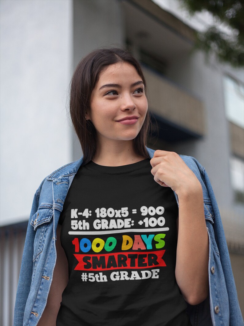 K&ouml;nnte beinhalten: Ein schwarzes T-Shirt mit dem Text "K-4: 180x5 = 900, 5th GRADE: +100, 1000 DAYS SMARTER, #5th GRADE" in Wei&szlig;, Rot, Gelb, Blau und Gr&uuml;n. Das T-Shirt wird von einer Person getragen, die eine Jeansjacke tr&auml;gt.