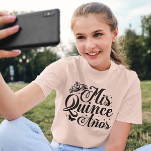 Puede incluir: Una persona joven lleva una camiseta rosa claro con el texto "Mis Quince Anos" en letra negra. La camiseta tiene una corona y estrellas. La persona se est&aacute; tomando una selfie con un tel&eacute;fono inteligente negro.