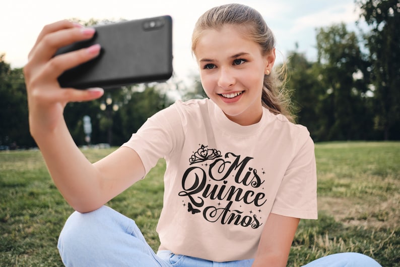 Puede incluir: Una persona joven lleva una camiseta rosa claro con el texto "Mis Quince Anos" en letra negra. La camiseta tiene una corona y estrellas. La persona se est&aacute; tomando una selfie con un tel&eacute;fono inteligente negro.