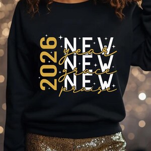 Peut inclure: Sweat-shirt noir avec l'inscription "2026 New Year New Grace New Praise" en or et blanc. Le motif comprend des &eacute;toiles, cr&eacute;ant un look festif et c&eacute;l&eacute;bratif.