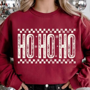 May include: Burgundy sweatshirt with the text "HO HO HO" in a distressed, off-white font. The text is framed by a checkered pattern. The design includes snowflakes and stars, creating a festive holiday theme.