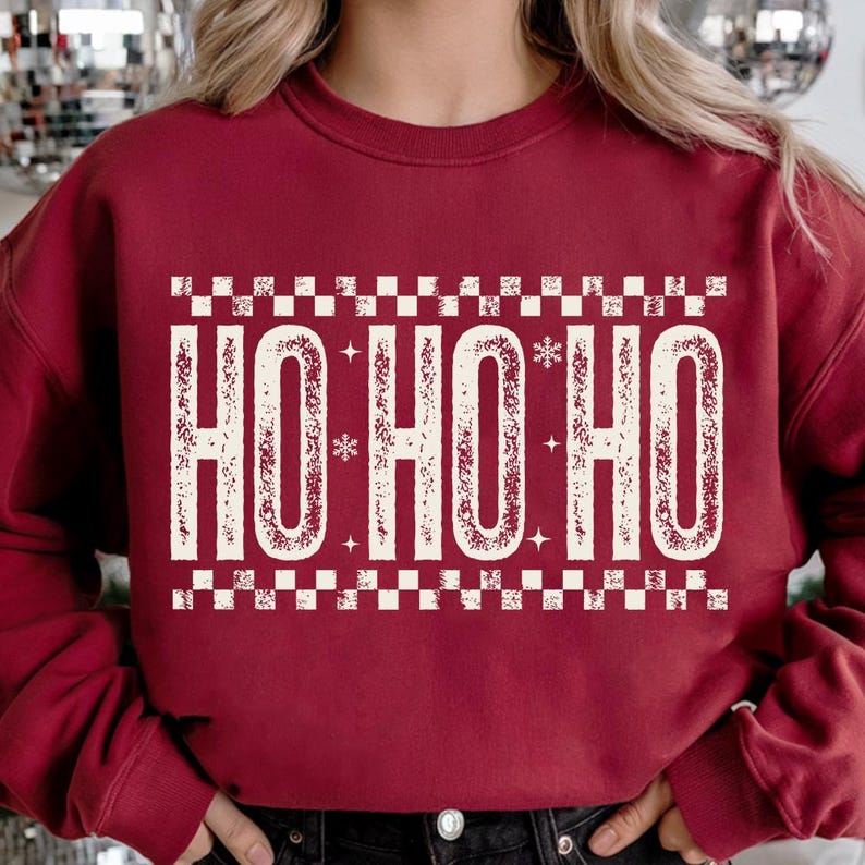 May include: Burgundy sweatshirt with the text "HO HO HO" in a distressed, off-white font. The text is framed by a checkered pattern. The design includes snowflakes and stars, creating a festive holiday theme.