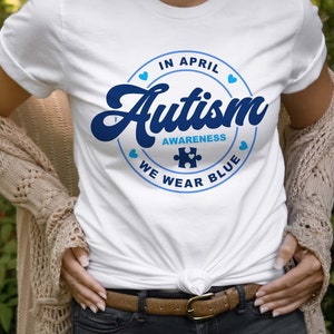 Puede incluir: Camiseta blanca con un gr&aacute;fico circular azul que dice "En abril, conciencia del autismo, usamos azul" con una pieza de rompecabezas azul en el centro.