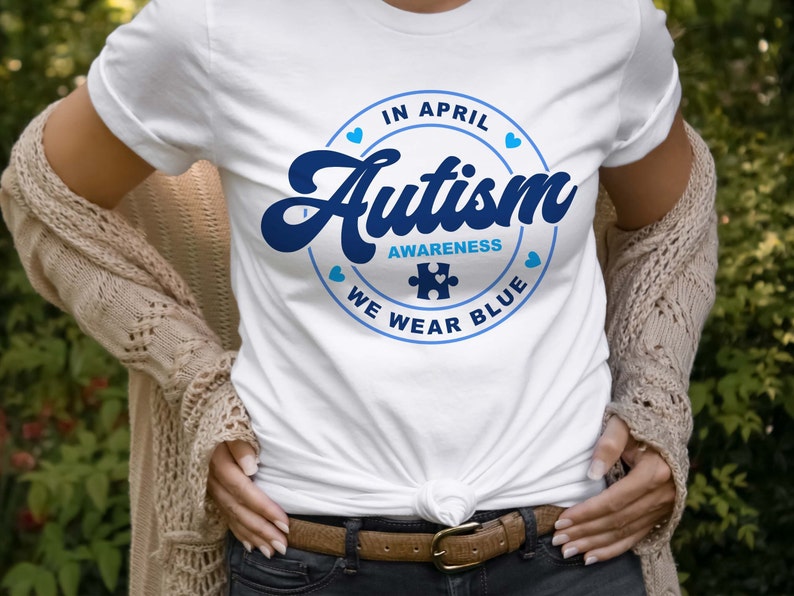 Puede incluir: Camiseta blanca con un gr&aacute;fico circular azul que dice "En abril, conciencia del autismo, usamos azul" con una pieza de rompecabezas azul en el centro.
