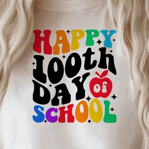 以下が含まれることがあります： 白いTシャツに、「Happy 100th Day of School」というカラフルなグラフィックがプリントされています。赤いリンゴと星も描かれています。