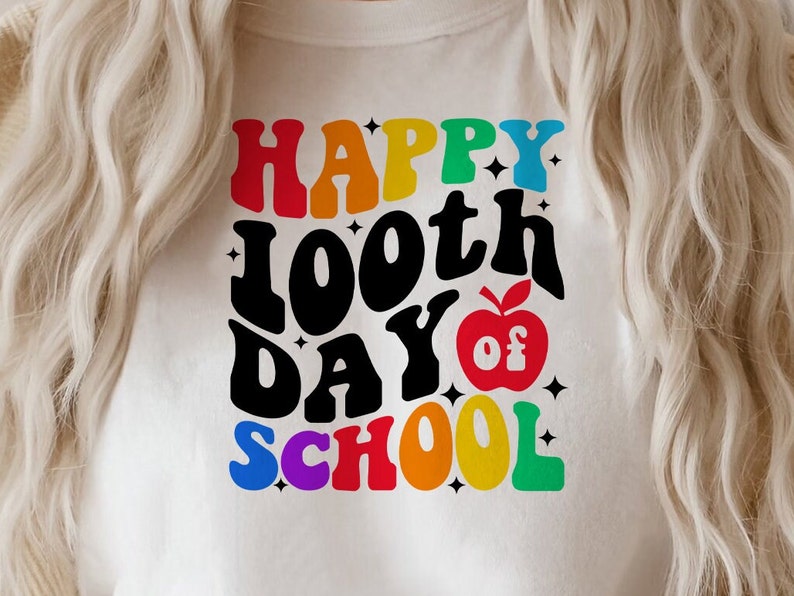 以下が含まれることがあります： 白いTシャツに、「Happy 100th Day of School」というカラフルなグラフィックがプリントされています。赤いリンゴと星も描かれています。
