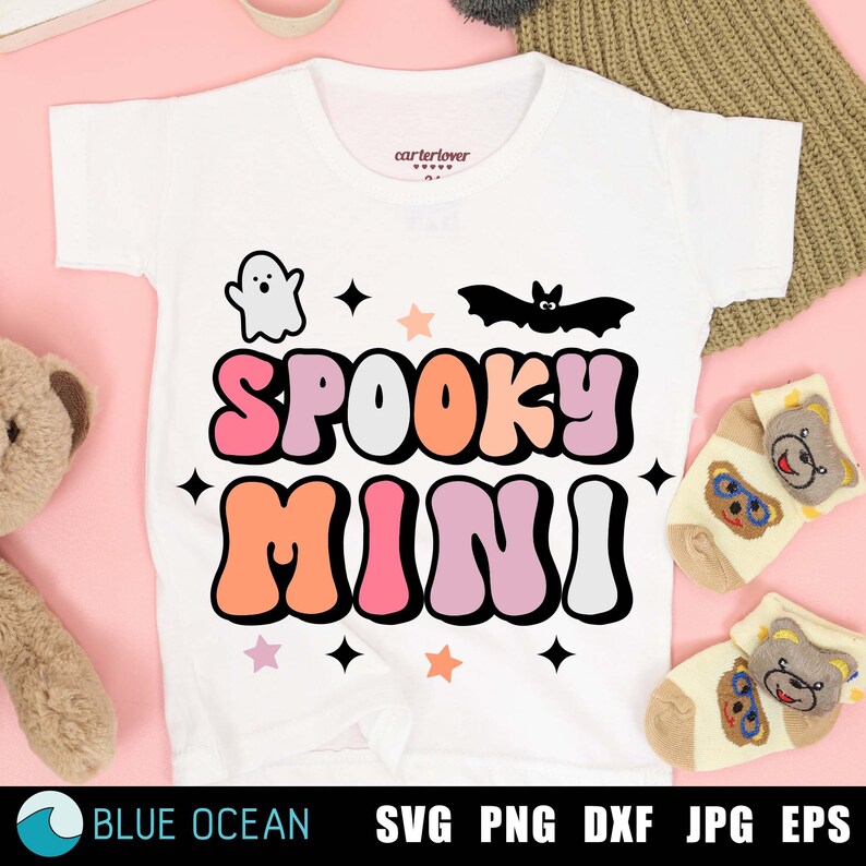 May include: White t-shirt with a graphic design of the words "Spooky Mini" in colorful, retro-style letters. The design also includes a ghost, a bat, and stars.