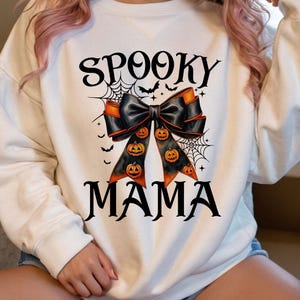 May include: White sweatshirt with a black bow with orange and black pumpkins and a spiderweb design. The text "Spooky Mama" is printed on the sweatshirt.
