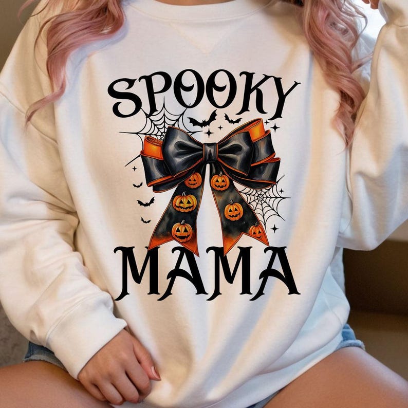 May include: White sweatshirt with a black bow with orange and black pumpkins and a spiderweb design. The text "Spooky Mama" is printed on the sweatshirt.