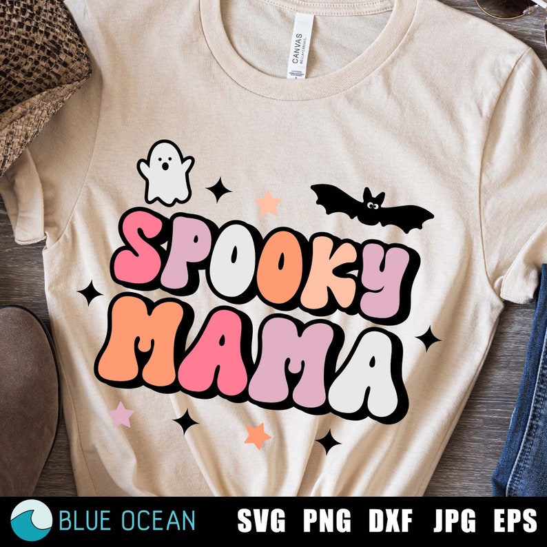 May include: A beige t-shirt with the text "Spooky Mama" in colorful, bubble-like letters. A ghost and a bat are above the text, and stars are scattered around the design.