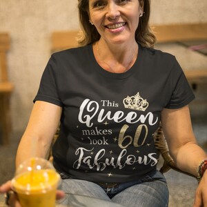 Op de afbeelding: Een vrouw draagt een zwart T-shirt met gouden glittertekst die "This Queen makes 60 look fabulous" zegt en een gouden kroonafbeelding. Ze houdt een doorzichtig plastic bekertje met sinaasappelsap vast.