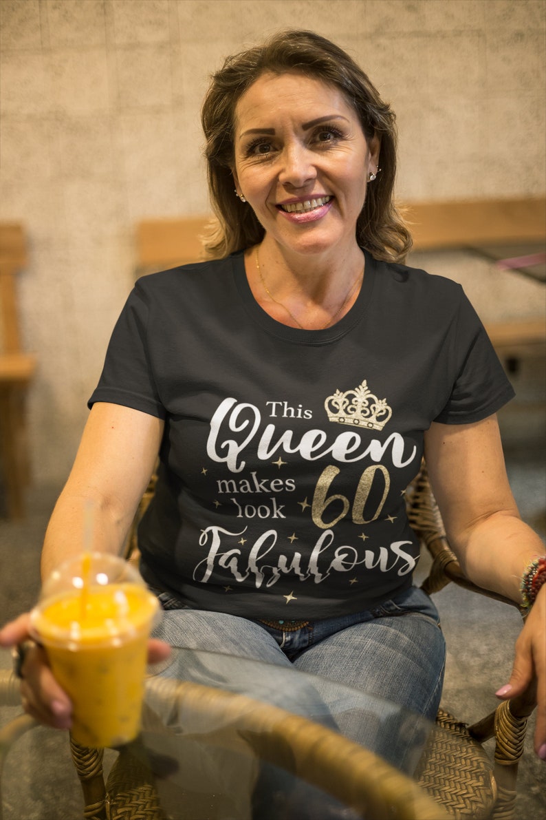 Op de afbeelding: Een vrouw draagt een zwart T-shirt met gouden glittertekst die "This Queen makes 60 look fabulous" zegt en een gouden kroonafbeelding. Ze houdt een doorzichtig plastic bekertje met sinaasappelsap vast.