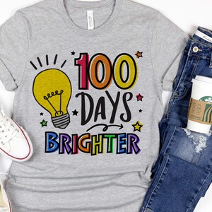 Peut inclure: Un t-shirt gris avec un graphique d'ampoule et le texte "100 Days Brighter" en couleurs arc-en-ciel. Le texte est soulign&eacute; en noir.