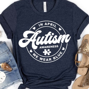 Puede incluir: Una camiseta azul marino con un gr&aacute;fico circular blanco que dice "En abril, conciencia del autismo, usamos azul" con una pieza de rompecabezas en el centro.