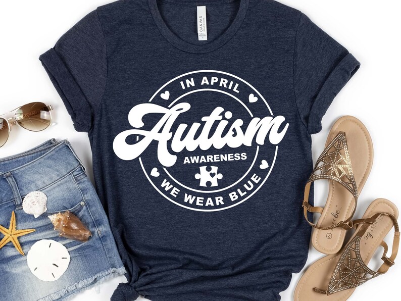 Puede incluir: Una camiseta azul marino con un gr&aacute;fico circular blanco que dice "En abril, conciencia del autismo, usamos azul" con una pieza de rompecabezas en el centro.
