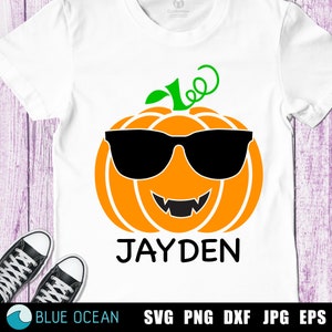 Op de afbeelding: Een wit T-shirt met een oranje pompoen die een zonnebril draagt en een brede glimlach laat zien. De pompoen heeft een groene steel en een zwarte omtrek. De tekst "JAYDEN" is onder de pompoen gedrukt.