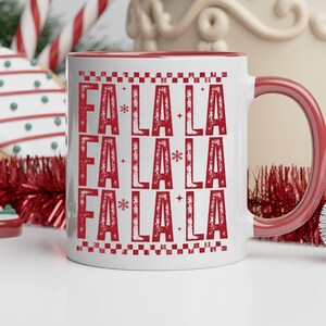 May include: White ceramic mug with a red interior and handle. The mug features the text "FA LA LA" in a repeating pattern in red, with snowflakes and star accents. Festive holiday theme.