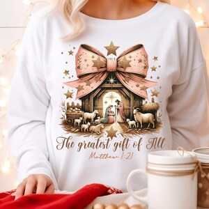 May include: White sweatshirt with a pink and gold bow and a nativity scene with the text "The greatest gift of all Matthew 1:21".