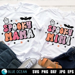 May include: Two white t-shirts with a Halloween theme. The larger shirt says "Spooky Mama" in pink, orange, and white letters with a ghost and bat graphic. The smaller shirt says "Spooky Mini" in the same colors with a ghost and bat graphic.