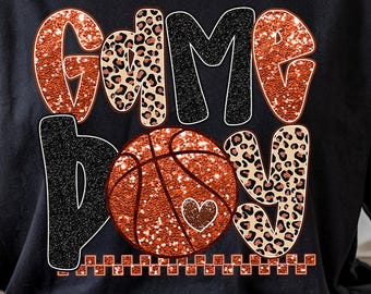 May include: Black t-shirt featuring the text "Game Boy" in orange glitter, black, and leopard print. A basketball with a heart is in the centre. The design incorporates a decorative border at the bottom of the graphic.