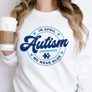 Puede incluir: Sudadera blanca con un gr&aacute;fico circular azul que dice "En abril, conciencia del autismo, usamos azul" con una pieza de rompecabezas azul en el centro.