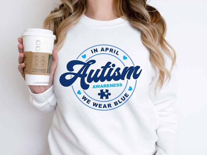 Puede incluir: Sudadera blanca con un gr&aacute;fico circular azul que dice "En abril, conciencia del autismo, usamos azul" con una pieza de rompecabezas azul en el centro.