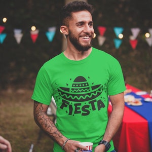 May include: A man wearing a green t-shirt with a black sombrero and the word "FIESTA" printed on it. He is smiling and holding a cup in his hand.