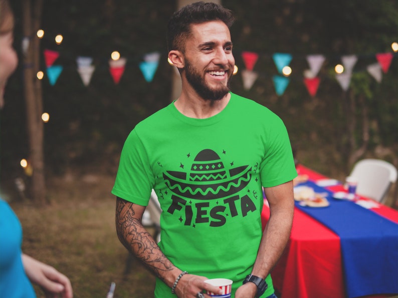 May include: A man wearing a green t-shirt with a black sombrero and the word "FIESTA" printed on it. He is smiling and holding a cup in his hand.