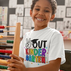 May include: A young boy wearing a white t-shirt with the text "OUT KINDERGARTEN" in rainbow colors and a graduation cap graphic. He is holding a wooden abacus.