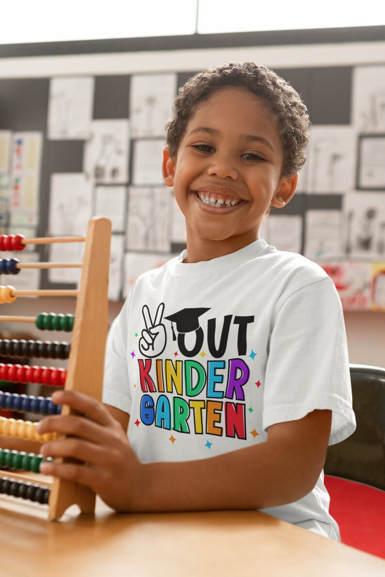 May include: A young boy wearing a white t-shirt with the text "OUT KINDERGARTEN" in rainbow colors and a graduation cap graphic. He is holding a wooden abacus.