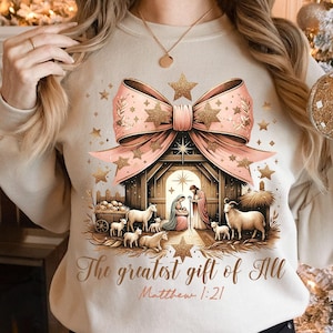 May include: A beige sweatshirt with a pink bow and gold glitter stars. The center of the design features a nativity scene with a wooden stable, Mary, Joseph, and baby Jesus. The text "The greatest gift of all" and "Matthew 1:21" is printed below the scene.