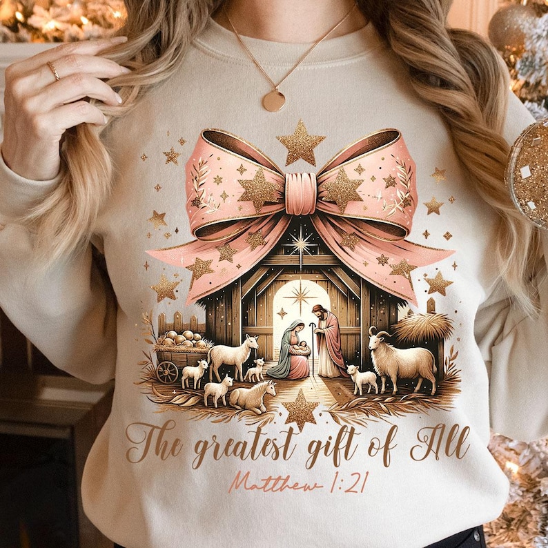 May include: A beige sweatshirt with a pink bow and gold glitter stars. The center of the design features a nativity scene with a wooden stable, Mary, Joseph, and baby Jesus. The text "The greatest gift of all" and "Matthew 1:21" is printed below the scene.