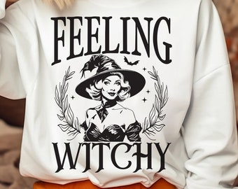 May include: A white sweatshirt with black text that reads "Feeling Witchy". A black and white illustration of a witch with a pointed hat and a bat flying above her head is in the center of the text.
