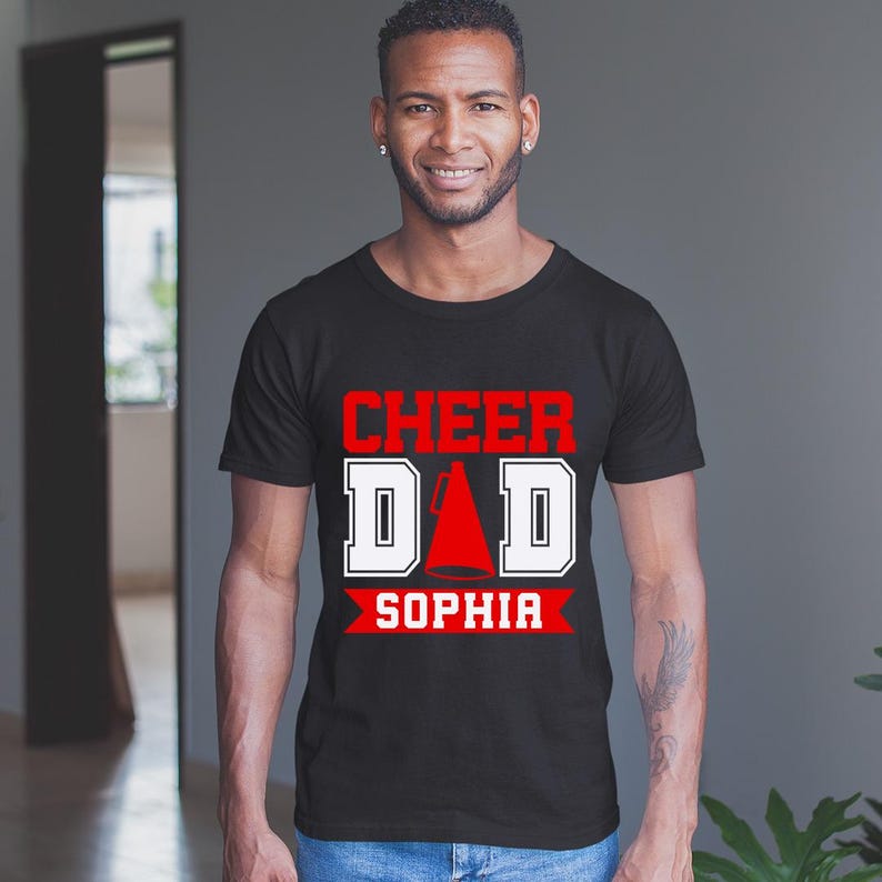 Pode incluir: Camiseta preta com texto branco que diz "Cheer Dad" com um gr&aacute;fico de megafone vermelho e o nome "Sophia" em vermelho abaixo.