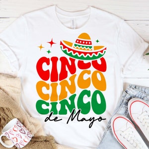May include: White t-shirt featuring a colourful graphic design that reads "Cinco Cinco Cinco de Mayo" with a sombrero on top.