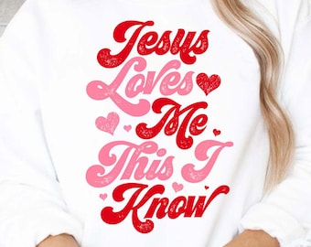 May include: A white sweatshirt with the text "Jesus Loves Me This I Know" in red and pink lettering with hearts. The text has a distressed, vintage look.