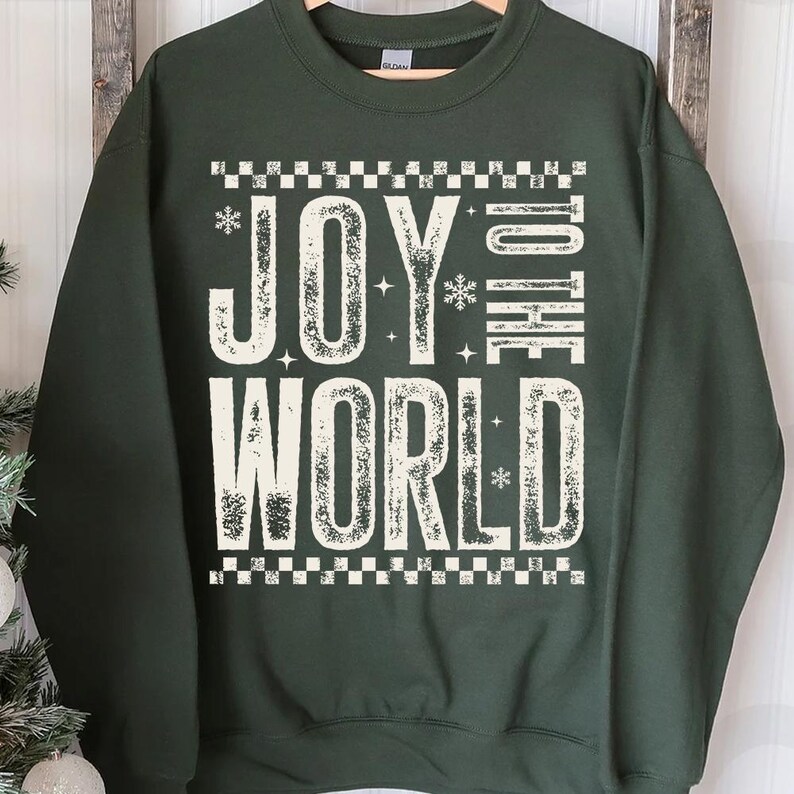 May include: Dark green sweatshirt with the text "JOY TO THE WORLD" in large, distressed white letters. The text is framed by a checkered pattern and snowflakes. A festive holiday-themed garment.