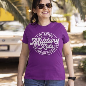 May include: A woman wearing a purple t-shirt with a white circle graphic that says "In April We Love Our Military Kids We Wear Purple".