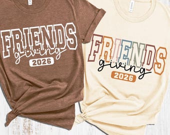 May include: Two short-sleeve crew neck t-shirts. The left shirt is brown with white text reading FRIENDS giving 2026. The right shirt is cream with multicolored text reading FRIENDS giving 2026. Both shirts feature a collegiate font style.
