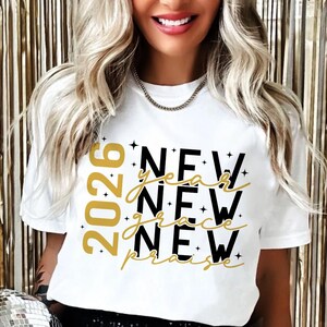 Peut inclure: T-shirt blanc avec l'inscription "2026 New Year New Grace New Praise" en noir et or. Le design comprend des &eacute;toiles. Le t-shirt a un col rond et des manches courtes.
