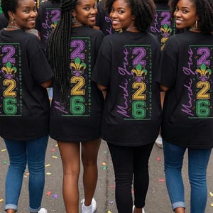 Op de afbeelding: Een groep mensen draagt zwarte t-shirts met een Mardi Gras-ontwerp. Het ontwerp bevat de tekst "2026" in paars, goud en groen, samen met de woorden "Mardi Gras" en "Let the Good Times Roll."