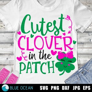 May include: A white t-shirt with green and pink text that reads "Cutest Clover in the Patch". A green four-leaf clover with a pink bow and a smiling face is to the right of the text.