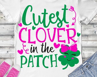 May include: A white t-shirt with green and pink text that reads "Cutest Clover in the Patch". A green four-leaf clover with a pink bow and a smiling face is to the right of the text.