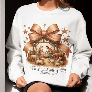 May include: White sweatshirt with a gold and brown illustration of the nativity scene. The illustration includes a bow, stars, and the text "The greatest gift of all Matthew 1:21".