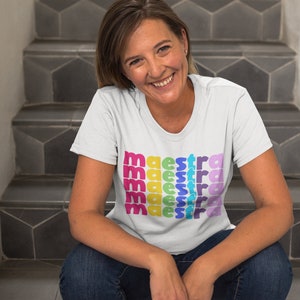 May include: A white t-shirt with the word "maestra" repeated in rainbow colours. The person wearing the shirt is sitting on a set of stairs.