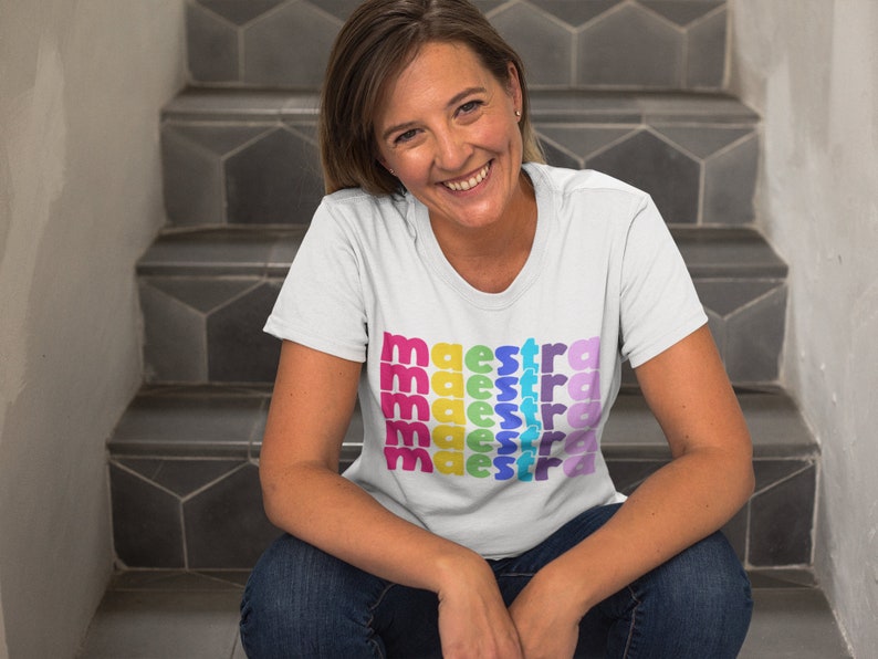 May include: A white t-shirt with the word "maestra" repeated in rainbow colours. The person wearing the shirt is sitting on a set of stairs.