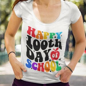 以下が含まれることがあります： 虹色のグラフィックがプリントされた白いTシャツ。グラフィックには「Happy 100th Day of School」の文字と赤いリンゴと星が描かれています。