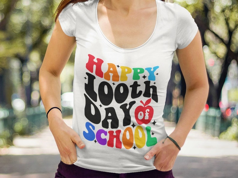 以下が含まれることがあります： 虹色のグラフィックがプリントされた白いTシャツ。グラフィックには「Happy 100th Day of School」の文字と赤いリンゴと星が描かれています。